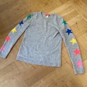 Charsmir tröja med rengbågiga stjärnor 🌟 - Säljer en grå charsmir tröja med färgglada stjärnor längs ärmarna. Tröjan har en rund halsringning och långa ärmar. Perfekt för att liva upp garderoben med lite färg!