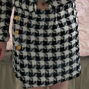 Rutig kjol från Stories - Snygg rutig kjol i svart och vit från Stories. Kjolen har en kort längd och är dekorerad med gula knappar på sidan. Perfekt för en stilren look.