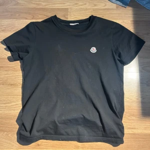 Svart t-shirt från Moncler - Säljer en stilren svart t-shirt från Moncler med korta ärmar och en liten broderad logga på bröstet. Perfekt för en casual look. T-shirten är i bomull och har en normal passform. Storlek 152 och den är köpt för 1300 kr på monclers hemsida.