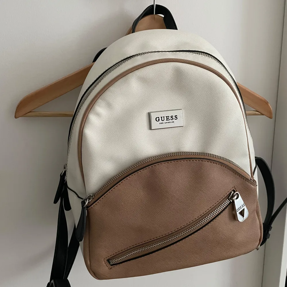 Snygg ryggsäck från Guess i beige och brunt med svarta axelremmar. Väskan har en praktisk dragkedja framtill och en stilren logotyp i silver. Perfekt för att bära dina dagliga nödvändigheter med stil.. Laukut & Käsilaukut.