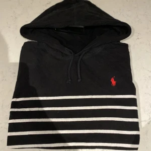 Svart och vit randig hoodie från Polo Ralph Lauren - Säljer en snygg svart hoodie från Polo Ralph Lauren med vita ränder och en röd logotyp på bröstet. 