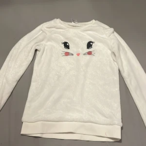 Vit långärmad tröja med kaninmotiv från Lindex - Säljer en söt vit långärmad tröja från Lindex med ett gulligt kaninansikte på framsidan. Tröjan har en mjuk och glittrig yta som ger en extra touch. Perfekt för den som älskar djurmotiv! 🐰✨