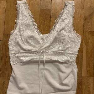Vit spetsprydd linnetopp - Säljer en vit linnetopp med vacker spetsdetalj och knytband framtill. Toppen har en omlott-design och är ärmlös, perfekt för varma dagar.
