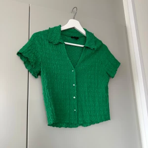 Grön kortärmad blus med knappar - Säljer en snygg grön blus med korta ärmar och knappar framtill. Blusen har en vågig textur och en snygg urringning. Perfekt för en färgglad look! Endast använd 2 gånger!