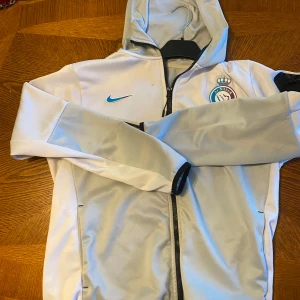 Vit träningsjacka från Nike - Säljer en vit träningsjacka från Nike med Al Nassr Football Club-logga. Jackan har en dragkedja framtill och en huva. Perfekt för fotbollsträning eller som en sportig vardagsjacka.