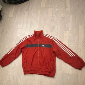 Röd vindjacka från Adidas - Säljer en röd vindjacka från Adidas med vita ränder längs ärmarna och en dragkedja framtill. Jackan har en hög krage och elastiska muddar vid ärmslut och nederkant. Perfekt för en sportig look!