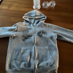 Grå hoodie med tryck - Säljer en grå hoodie med dragkedja och ett unikt tryck på framsidan. Tröjan har långa ärmar och en praktisk huva. Perfekt för en avslappnad stil. Det är av märket aim for the moon. Möjligtvis att några pärlor fallit bort men de inte så man lägger märke till det så mycket. 