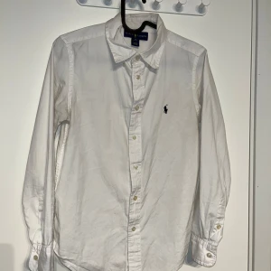 Vit skjorta från Ralph Lauren - Säljer en klassisk vit skjorta från Ralph Lauren med broderad logga på bröstet. Skjortan har långa ärmar och knappar framtill. Perfekt för en stilren look. Lite liten i storleken passar till 12-13.