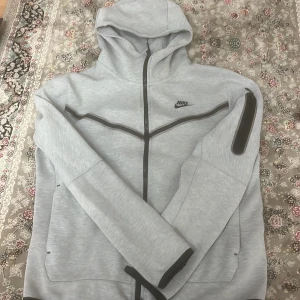 Grå huvtröja från Nike - Säljer en stilren grå huvtröja från Nike med dragkedja och huva. Tröjan har långa ärmar och en ficka med dragkedja på ärmen. Perfekt för en sportig look eller avslappnade dagar.