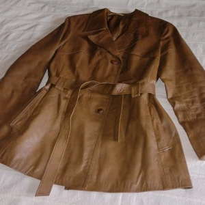 Vintage brun/tanfärgad blazer/jacka/skinnjacka med bälte - Svinsnygg brun skinnjacka/lite längre blazer med bälte och knappar, från Tjäders Malung (originalplagg) i äkta getskinn. Några märken i lädret och den är lite småfläckig invändigt. Storlek 40 men passar fler storlekar också, beroende på hur man vill ha den.