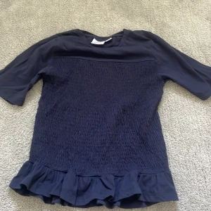 Mörkblå blus med volang från Gina Tricot - Supersöt mörkblå blus från Gina Tricot med smockad front och baksida. Toppen har korta ärmar och en bred volang längst ner som ger en extra touch. Perfekt för dig som gillar en stilren men ändå lekfull look.