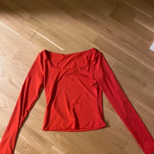 Röd långärmad topp med vid urringning - Säljer en snygg röd långärmad topp med vid, nästan fyrkantig urringning. Modellen är figurnära och enkel, perfekt att styla med jeans eller kjol. Tunt och mjukt material som känns skönt mot huden.