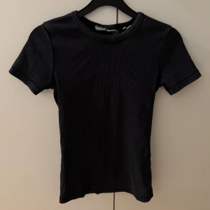 Svart ribbad t-shirt - En svart ribbad t-shirt med rund hals och korta ärmar. T-shirten har en enkel och clean look, sitter snyggt och är lätt att matcha till olika outfits. Perfekt basplagg för garderoben.