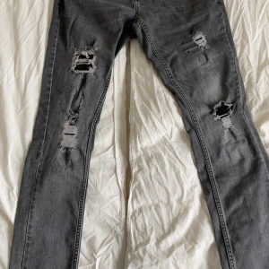Grå slitna jeans från Tazzio Fashion - Stilrena och moderna jeans från Tazzio Fashion i grå tvätt med kraftiga slitningar för en trendig och avslappnad look. Jeansen har en smal passform som sitter snyggt och ger en tuff streetwear-stil.  👖 Färg: Mörkgrå tvätt 🧵 Design: Distressed/ripped med slitna detaljer på båda benen 👟 Passar perfekt till sneakers och t-shirt eller hoodie 📏 Storlek: W/32 L/32 📦 Skick: Begagnade men i fint skick – designade slitningar, inga skador utöver det  Perfekta jeans för dig som vill sticka ut lite extra!