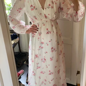 Vit långklänning med blommigt mönster - Superfin långklänning i vit med rosa och lila blommor, v-ringad och med långa puffärmar. Klänningen har ett luftigt fall och är tillverkad i ett lätt, strukturerat tyg som känns perfekt för sommaren. 