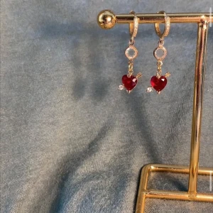 Guldiga örhängen med röda hjärtan - Supersöta örhängen i guldton med hängande röda hjärtan och små gnistrande stenar. Hjärtana har en pil-detalj som ger en lekfull och romantisk vibe. Perfekta för att lyfta vilken outfit som helst med lite extra kärlek.