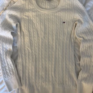 Sweatshirt från Tommy hilfigur  - Vit kabelstickad tröja från Tommy Hilfiger med rund halsringning och diskret logga broderad på bröstet. Långärmad modell i mjuk bomull, perfekt för lager-på-lager och snygg till jeans eller kjol.