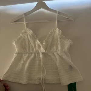 Vit linnetopp med volang och snörning - Supersöt vit linnetopp med smala axelband, volangkant vid bysten och snörning framtill. Toppen har en lätt strukturerad yta och är perfekt för varma dagar. Den har en luftig passform och markerad midja för en feminin look.