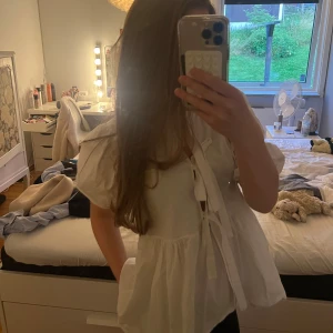 Vit blus med puffärm och knyt - Supersöt vit blus med puffiga korta ärmar och knyt framtill. Modellen är luftig och har en lätt volang nedtill som ger en romantisk vibe. Perfekt för dig som gillar en feminin och trendig look. Tillverkad i mjuk bomull som känns skön mot huden. Skriv för fler bilder!