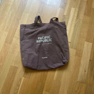 Brun tygväska  från Pull&Bear - En brun shoppingväska från Pull&Bear med trycket 'Pacific Republic Teen Department' på framsidan. Väskan är i bomull och har långa handtag som gör den enkel att bära på axeln. Perfekt för att slänga ner allt du behöver till skolan eller stan.