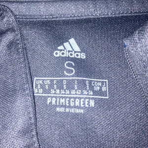 Svart Adidas half zip träningströja S - Svart långärmad träningströja från Adidas med klassiska vita ränder längs ärmarna och halv dragkedja framtill. Tillverkad i funktionsmaterialet Primegreen, perfekt för sport eller gym. Adidas-logga på bröstet och tight passform.