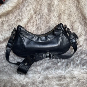 Svart axelväska i skinn med dragkedja - Snygg svart axelväska i skinn med justerbart axelband och guldfärgad dragkedja. Väskan har en rundad form och fina sömdetaljer framtill. Perfekt storlek för det viktigaste och passar till många olika outfits.