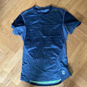 Nike Pro Combat blå träningströja - Nike Pro Combat Dri-Fit t-shirt i grått med subtila ränder och coolt mönster i gult på ryggen. Kortärmad och tillverkad i lätt, ventilerande polyester som passar perfekt för intensiva träningspass. Tight passform och sportig look med logga på bröstet och nederkanten.