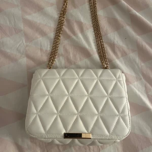Vit quiltad axelväska med guldkedja - Snygg vit axelväska med quiltat mönster och guldfärgad kedja som axelrem. Väskan har en stilren rektangulär form och stängs med ett metallspänne framtill. Perfekt för att lyfta vilken outfit som helst.