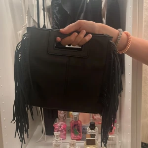 Svart handväska med fransar - Snygg svart handväska i skinnimitation med rektangulärt handtag och långa fransar på sidorna. Väskan har en stilren design och är rymlig inuti, perfekt för att bära med sig det viktigaste. Passar dig som gillar trendiga detaljer och en edgy look.