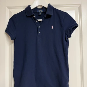 Marinblå pikétröja från Ralph Lauren - Klassisk marinblå pikétröja från Ralph Lauren med korta ärmar och krage. Tröjan har knäppning framtill med tre knappar och den ikoniska rosa broderade hästloggan på bröstet. Tillverkad i mjuk bomull, perfekt för en clean och sportig look.