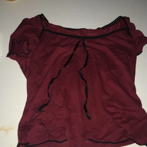 Vinröd blus med svarta spetsdetaljer - Säljer en vinröd blus med korta puffärmar och svarta spetsdetaljer längs halsringningen. Blusen har en söt knytning framtill och är lätt och luftig i materialet. Perfekt för dig som gillar romatiska plagg och feminina plagg.