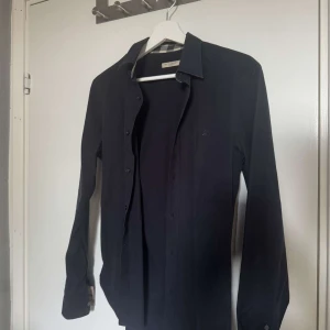 Svart skjorta från Burberry  - Stilren svart skjorta från Burberry i bomull med klassisk krage och knappar framtill. Insidan av manschetterna och kragen har Burberrys ikoniska rutiga mönster i beige och svart. Pris ej hugget i sten!