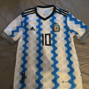 Argentina Messi #10 Adidas fotbollströja - Säljer en officiell Argentina fotbollströja från Adidas med Messi och nummer 10 på ryggen. Tröjan har ett unikt blått och vitt mönster, svarta detaljer och klassiska Adidas-ränder på axlar och ärmslut. Tillverkad i lätt och ventilerande material, perfekt för match eller träning.