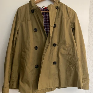 Trenchcoat  - Snygg trenchcoat med dubbelknäppning och huva. Jackan har långa ärmar med knappar vid ärmslut, två fickor fram och ett rutigt innerfoder. Perfekt för dig som gillar klassisk stil med en twist💗
