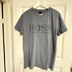 Grå Hugo Boss t-shirt med logga - Snygg mörkgrå t-shirt från Hugo Boss med stor BOSS-logga framtill. Klassisk rund halsringning och korta ärmar. Tillverkad i mjuk bomull som känns skön mot huden. Perfekt för dig som gillar stilrena och trendiga plagg med premiumkänsla.