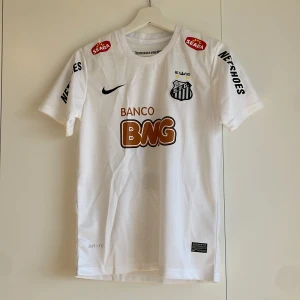 Santos Neymar Jr #11 Nike fotbollströja - Vit Santos fotbollströja från Nike med Neymar Jr och nummer 11 på ryggen. Klassisk design med klubbmärke, sponsorer och svarta detaljer. Tillverkad i lätt, ventilerande Dri-FIT-material. Korta ärmar och rund hals. Matchande shorts med nummer 11 och klubbmärke ingår.