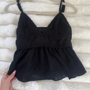 Svart ärmlös linnetopp med spetsdetaljer - Supersöt svart linnetopp med tunna axelband och v-ringning. Toppen har broderade prickar i tyget och fina spetsdetaljer under bysten. Den är lätt utsvängd nertill och har en luftig känsla, perfekt för varma dagar.