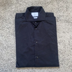 Svart skjorta från Bläck, tailored fit - Klassisk svart skjorta från Bläck i storlek S, tailored fit. Skjortan har knappar framtill och en stilren krage. Perfekt för dig som gillar en clean och enkel look. Materialet känns mjukt och följsamt mot huden.