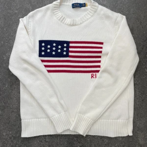 Polo Ralph Lauren tröja  - Vit stickad tröja från Polo Ralph Lauren med amerikansk flagga i blått, rött och vitt på bröstet. Klassisk rund halsringning och ribbade muddar. Perfekt för dig som gillar preppy stil och vill sticka ut med en ikonisk detalj. Priset är ej fast så kom med bud! 😁