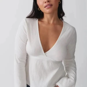 Vit omlottopp med knytband - Säljer en vit omlottopp med lång ärm och djup v-ringning. Toppen har en snygg knytning i ryggen och är gjord i ett lätt, strukturerat tyg som ger en fräsch känsla. Perfekt att styla med jeans eller kjol för en clean look.