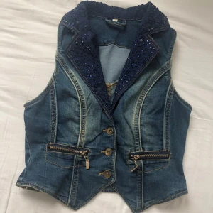 Jeansväst med spets och paljetter - Snygg jeansväst/topp i blå denim med cool spetskrage dekorerad med mörkblå paljetter. Västen har markerade sömmar, tre knappar framtill och två fickor med dragkedja. Perfekt för fest och för att ge din outfit något extra. Tryck gärna på KÖP NU, pris kan diskuteras💕