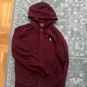 Vinröd hoodie från Polo Ralph Lauren - Snygg vinröd hoodie från Polo Ralph Lauren med dragkedja och huva. Klassisk logga broderad på bröstet och praktiska fickor framtill. Perfekt för chill dagar eller när du vill ha en stilren streetlook.