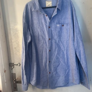 Blå skjorta - Snygg ljus blå skjorta från Homebound i storlek XXL. Skjortan har klassisk krage, bröstficka med broderad logga och bruna knappar. Perfekt för dig som gillar en enkel och stilren look nu i sommar 