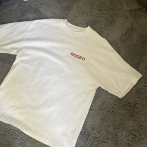 Vit RODEO t-shirt från Bolaget - Vit t-shirt från Bolaget med röd RODEO-text på bröstet och stor Vegas-inspirerad print på ryggen. Klassisk passform, rund hals och korta ärmar. Perfekt statement-plagg för dig som gillar unika tryck och streetstyle. Denna säljs endast på konserter.