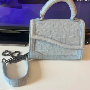 Ljusblå jeans axelväska från Zara - Snygg ljusblå axelväska från Zara i jeansmaterial med croco-mönstrad struktur. Väskan har ett kort handtag upptill och en lång kedjerem för att bära över axeln. Magnetlås och metallfötter undertill ger extra detaljerad look.