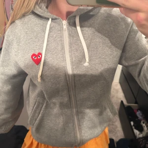 Grå fleecehoodie från play - Grå zip-hoodie från Play med det ikoniska röda hjärtat broderat på bröstet. Jackan har huva med vita snören, dragkedja framtill och fickor på sidorna. Perfekt för en chill och stilren look.