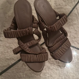 Bruna sandaler med klack från Burberry - Unika bruna sandaler från Burberry med höga klackar och flera vadderade remmar i skinn. Snyggt rynkade detaljer på remmarna ger en cool och trendig look. Perfekta för dig som vill sticka ut med ett exklusivt statement.