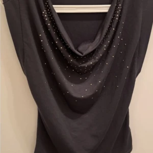 Grå topp med silver detaljer från Guess - Grå topp från Guess med snygg draperad halsringning och små silver färgade nitar framtill. Toppen är ärmlös och har en loose passform upptill perfekt för dig som gillar en stilren look med lite edge. Materialet känns mjukt och följsamt mot kroppen.