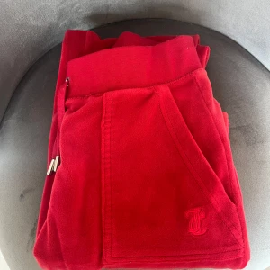 Röda mjukisbyxor från Juicy Couture - Riktigt snygga röda mjukisbyxor från Juicy Couture i mjuk velour. Byxorna har dragsko i midjan, fickor fram och bak med silverfärgade knappar och broderad JC-logga. Perfekta för en chill och trendig look. Använda cirka 3-5 gånger bara.