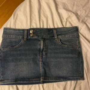 Blå jeanskjol med fickor - Snygg blå jeanskjol i klassisk modell med två framfickor, två bakfickor och dubbla knappar i midjan. Kjolen är kort och har bälteshällor, perfekt till en avslappnad stil. Materialet är denim och färgen är mellanblå.